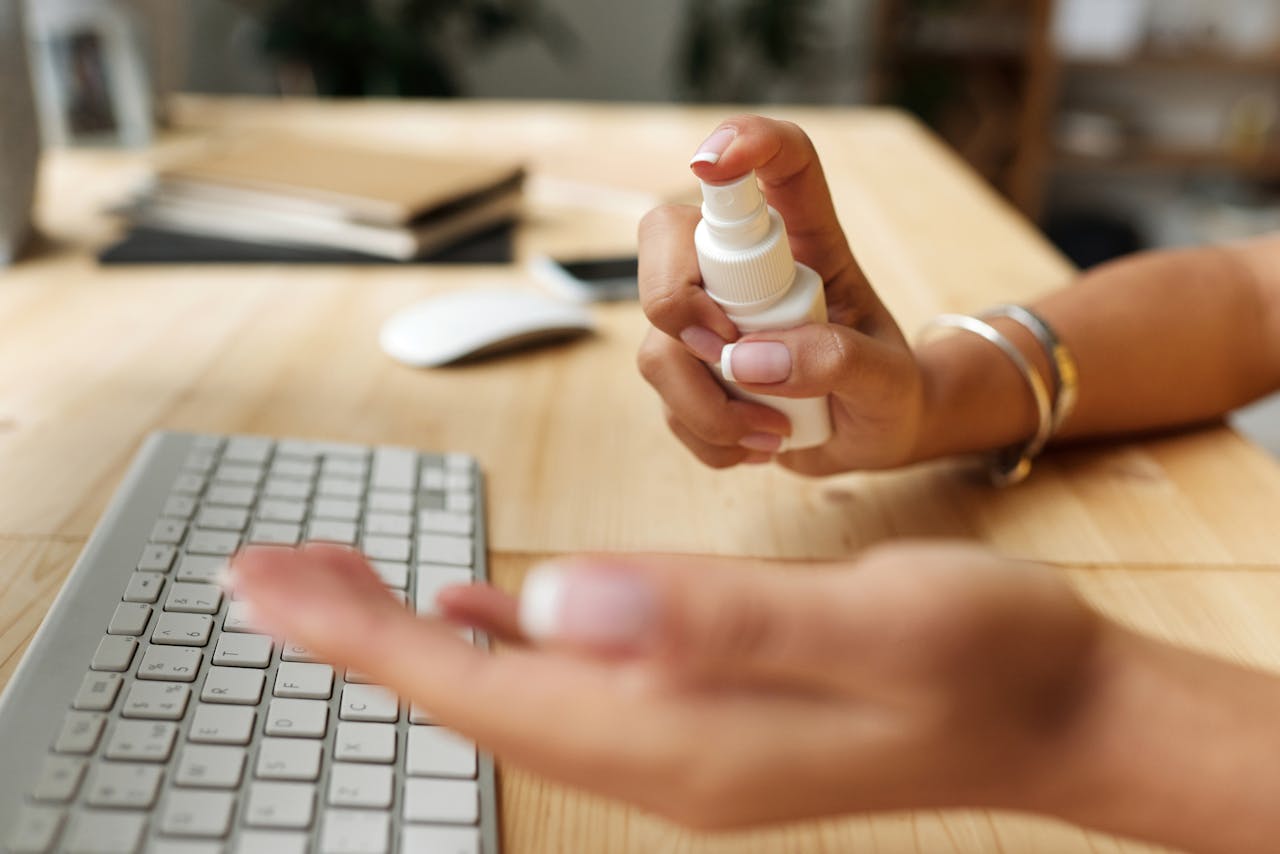 Person sanitizing hands at a desk with spray bottle, keyboard, and computer mouse.